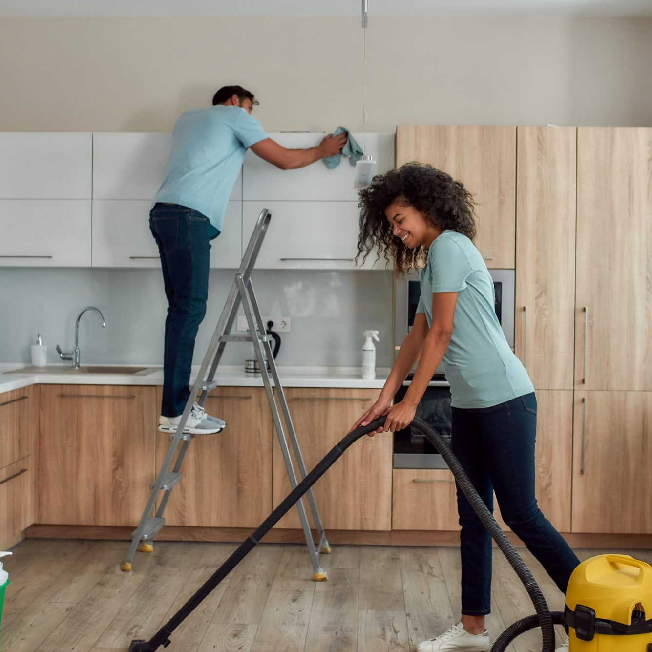 Cleaning experts. Two young professional cleaners in uniform working together in the kitchen. Young caucasian man. Young caucasian men cleaning modern kitchen furniture, smiling afro american women using vacuum cleaner. Cleaning services.
